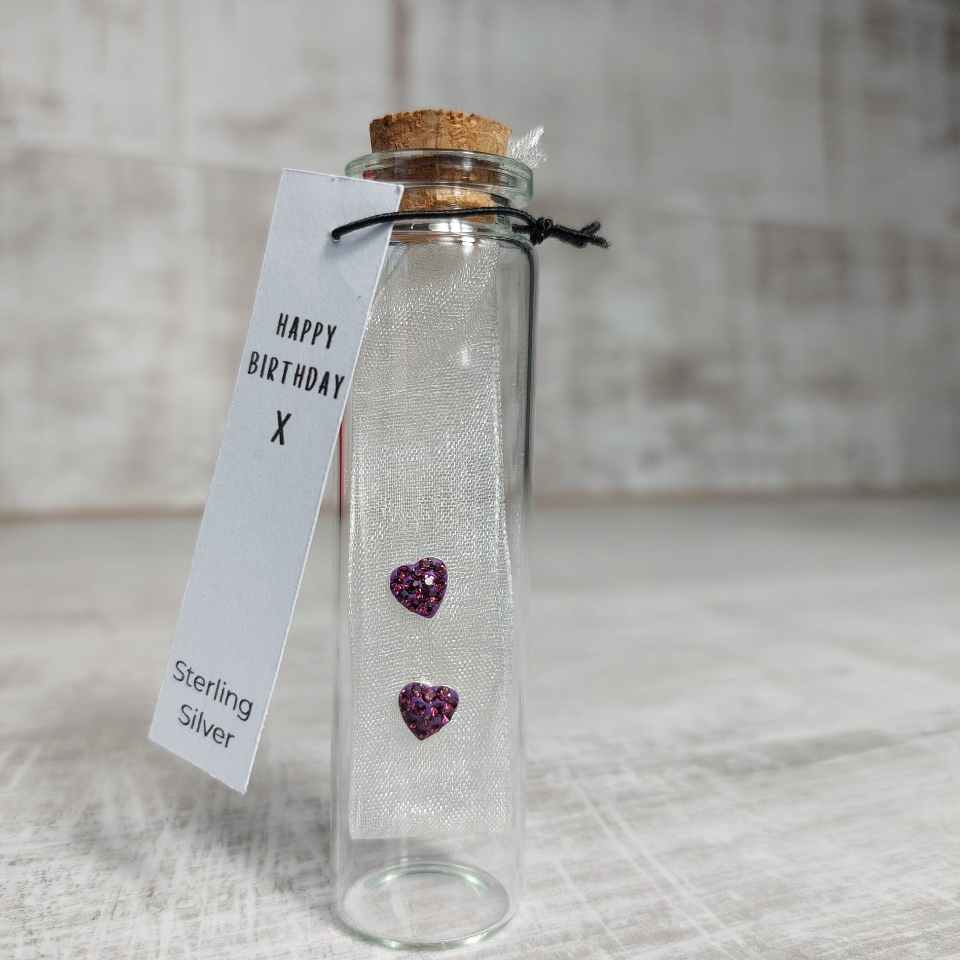 Pair of purple amethyst earrings on a fabric background with a printed tag saying Happy Birthday and sterling silver. Earrings are presented in a small bottle with cork stopper.