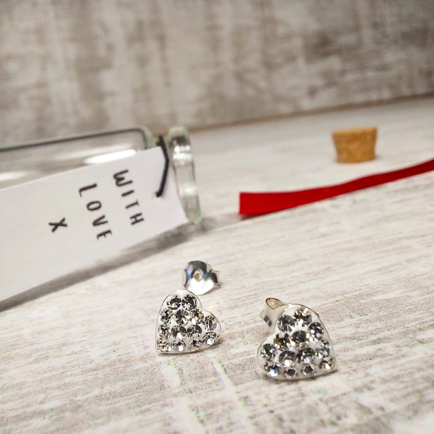 Pair of heart earrings with tiny crystals all over creating sparkle. Pictured with a glass bottle and red ribbon in the background.