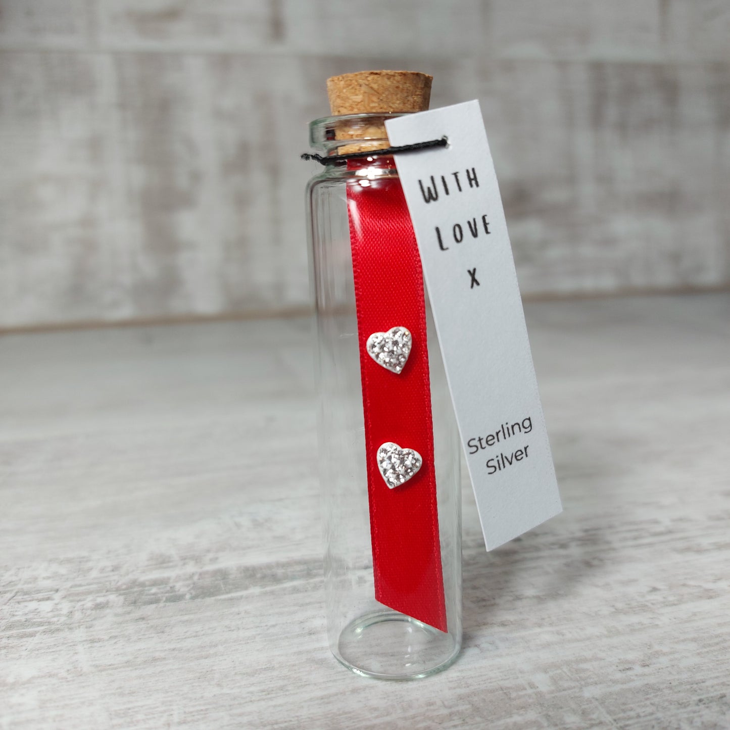 Pair of heart shaped crystal earrings on a red ribbon. These are presented in a cork topped glass bottle and have a gift tag attached. The gift tag has the words With Love and  sterling silver.