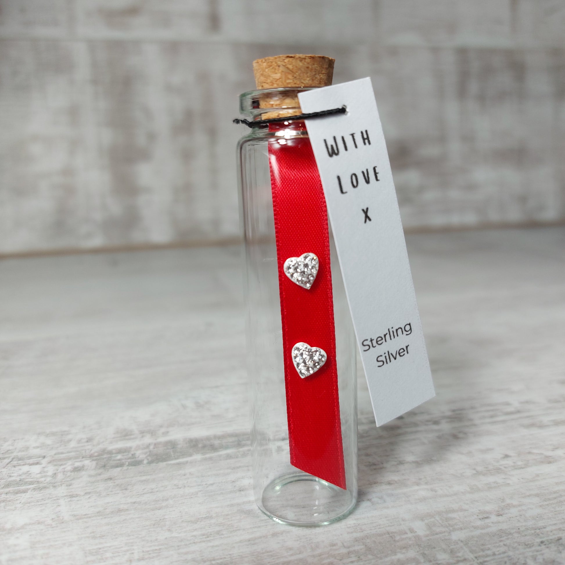 Pair of heart shaped crystal earrings on a red ribbon. These are presented in a cork topped glass bottle and have a gift tag attached. The gift tag has the words With Love and  sterling silver.