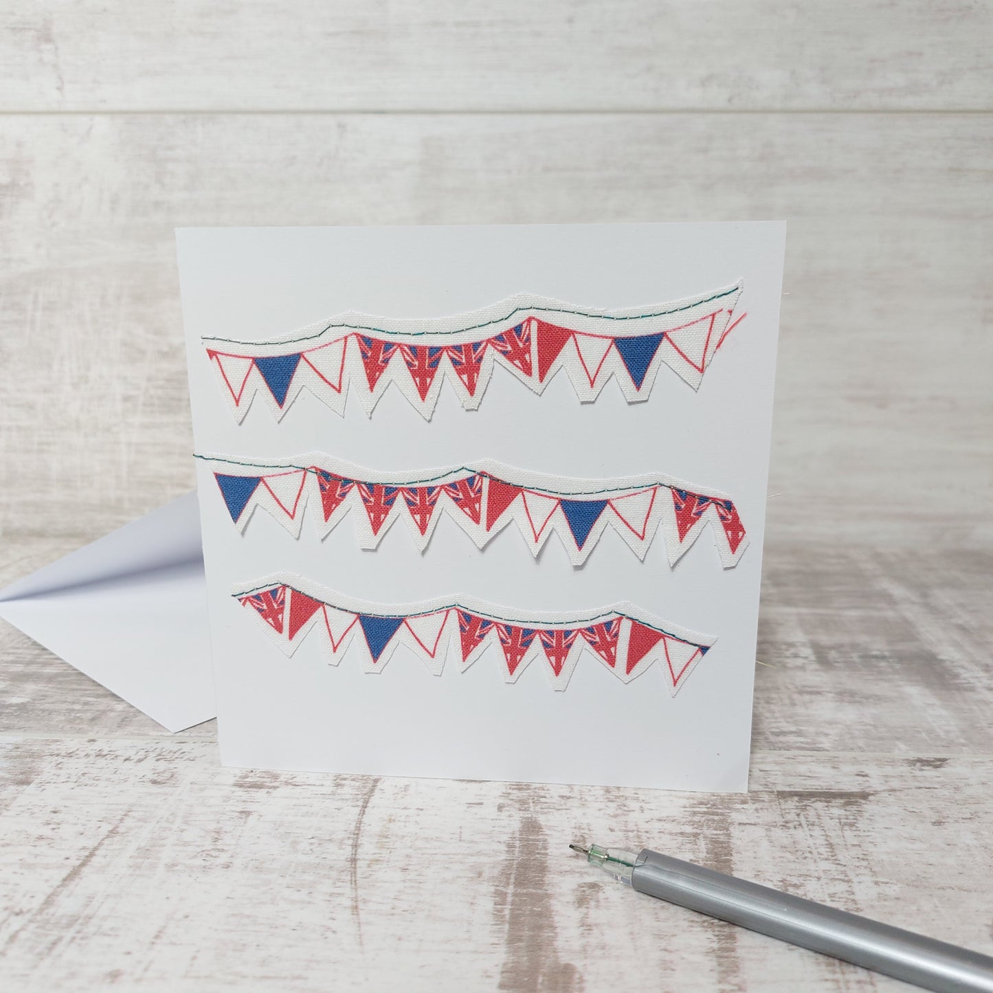 Square greetings card of red, white and blue bunting stitched onto it.  3 rows with multiple bunting with union jacks on. 