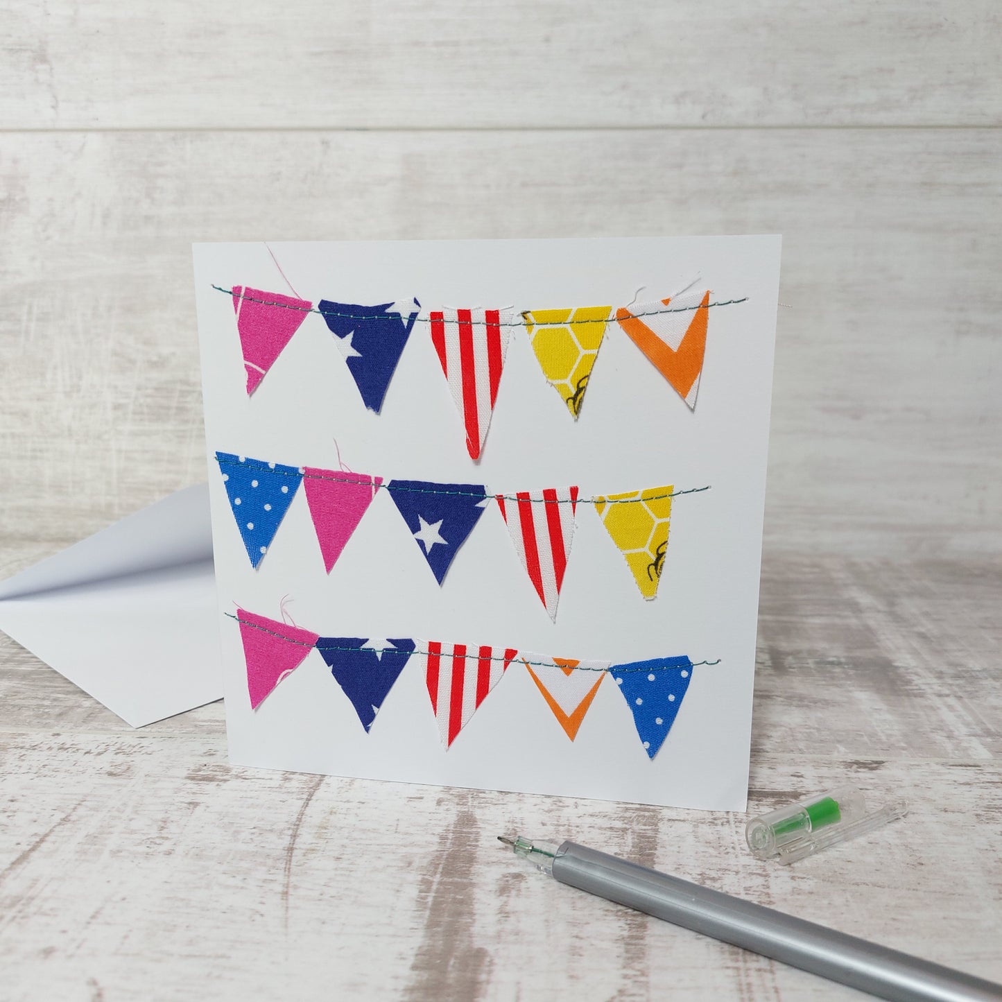 Square greetings card with 3 rows of multi coloured bunting stitched onto it. Each row has 5 triangle pieces of fabric bunting. 